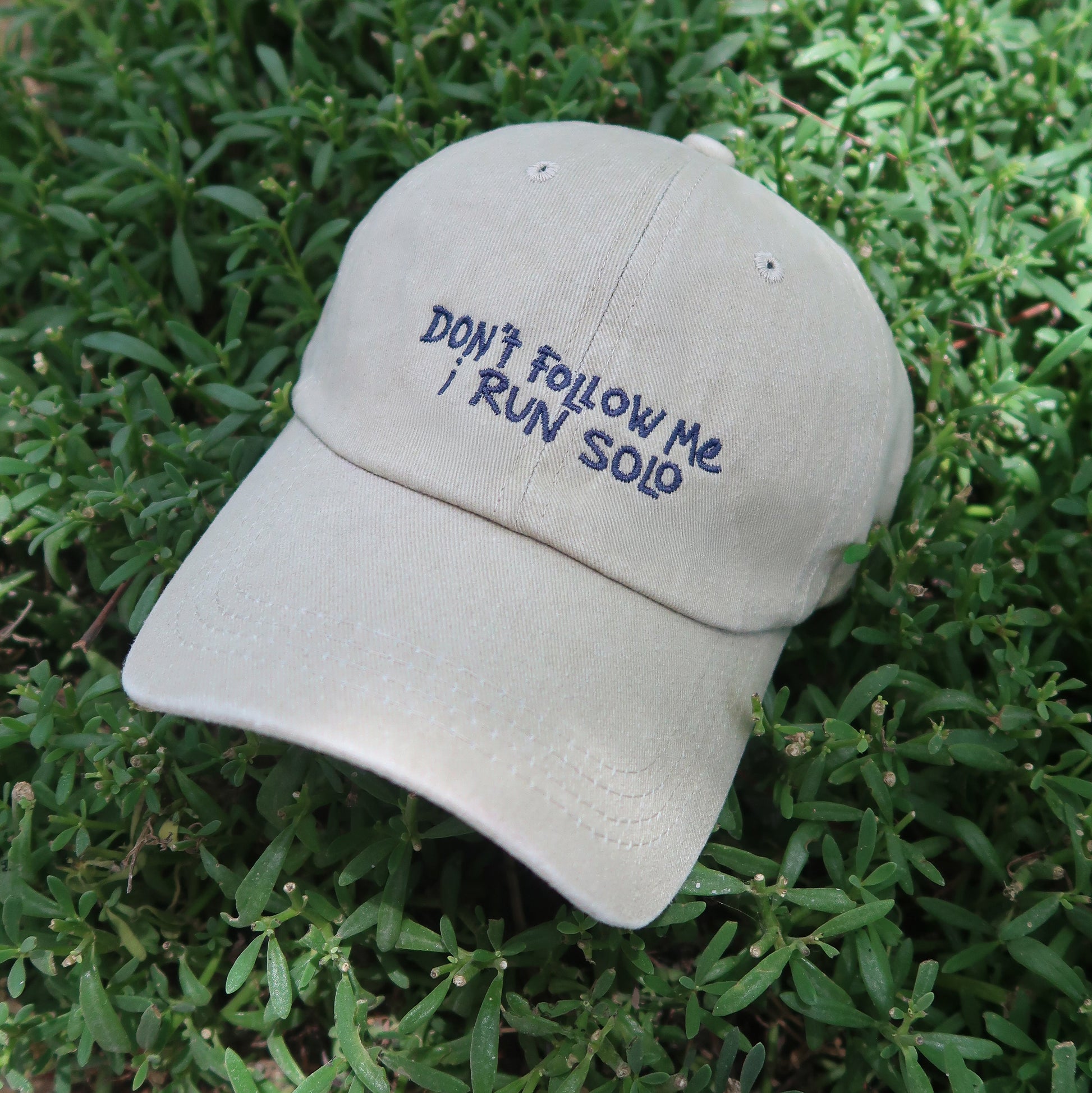 Beige cap with 'Don't follow me, I run solo' text on green grass